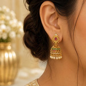 Könnte beinhalten: Ein goldener Jhumka-Ohrring mit einem verzierten Stecker, einem tropfenförmigen rosa Stein, einer glockenförmigen Kuppel mit grünen und rosa Steinen, klaren Kristallen und einem unteren Rand aus kleinen weißen Perlen, die an der Basis hängen.