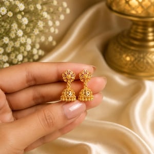 May include: A pair of gold-toned jhumka earrings featuring a delicate floral stud top connected to a bell-shaped drop. The bell component displays intricate filigree metalwork and is finished with a row of small, dangling gold beads at the base.
