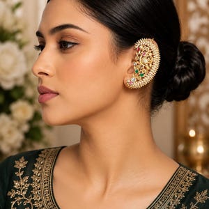 May include: A gold ear cuff featuring an intricate filigree design adorned with small white pearls along the outer edge and accented with embedded red, green, and clear stones. The piece follows the curve of the ear, showcasing detailed metalwork.