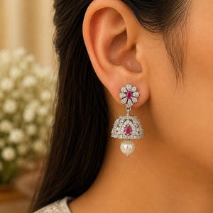 May include: A silver-toned jhumka earring featuring a flower-shaped stud with a central teardrop-shaped pink stone surrounded by clear crystals. The bell-shaped drop is encrusted with clear crystals, a pink teardrop stone, and a white pearl bead.
