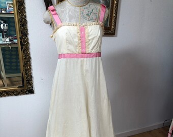 Könnte beinhalten: Ein langes weißes Kleid mit rosa Besatz und Spitzendetails. Das Kleid hat ein tailliertes Oberteil und einen fließenden Rock.