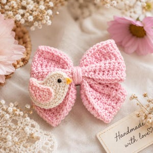 Può includere: Un fiocco rosa all'uncinetto fatto a mano con una piccola applicazione di uccello. L'uccello è color crema e rosa con un becco giallo e un occhio nero. Il fiocco è su una superficie bianca con fiori e un'etichetta che dice "Handmade with love".
