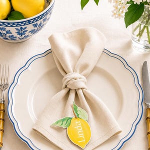 Puede incluir: Una servilleta de tela beige anudada, con una tarjeta de sitio en forma de limón con el nombre Linda impreso. La servilleta descansa sobre un plato blanco con borde festoneado y una línea azul, flanqueada por un tenedor y un cuchillo de plata.