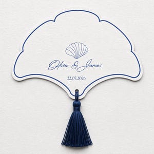 May include: A white fan-shaped card with a dark blue border, featuring a blue seashell illustration, the text Olivia & James in script, and the date 22.07.2026. A dark blue tassel is attached to the bottom center of the card through a small hole.