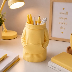 May include: A yellow pencil holder shaped like a miniature hooded sweatshirt with a ribbed collar, ribbed hem, and a front kangaroo pocket. The container is filled with several pens and pencils, standing upright as a functional desk accessory.