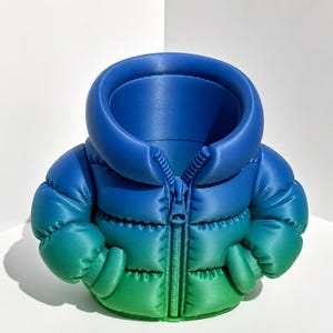 May include: A desk organiser shaped like a miniature puffer jacket with a gradient colour scheme transitioning from deep blue at the collar to a vibrant green at the base. The item features a central zip detail and a hollow interior opening.