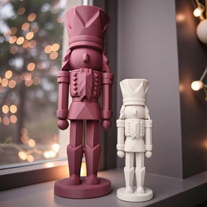 May include: Two geometric nutcracker figurines standing upright. The larger nutcracker is a deep maroon colour, while the smaller nutcracker is a solid off-white. Both figures feature faceted, angular surfaces and stand on circular bases.