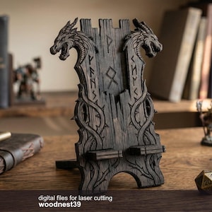 Puede incluir: Un soporte de teléfono de madera oscura con tallas de cabeza de dragón y símbolos rúnicos. El soporte está sobre un escritorio de madera con una libreta de cuero, un bolígrafo y otros objetos decorativos. El texto "digital files for laser cutting woodnest39" está en la parte inferior.