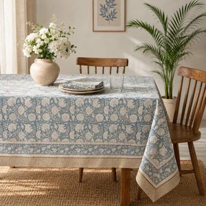 Puede incluir: Un mantel rectangular con un estampado floral en azul y blanco realizado mediante bloques de impresión y un borde a rayas. El mantel cubre una mesa de madera, sobre la cual hay un jarrón de cerámica con flores blancas y servilletas.