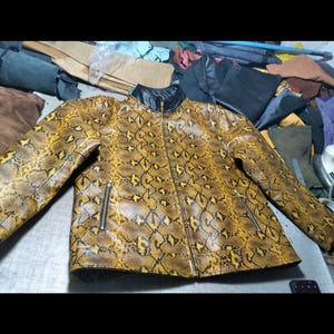 May include: A long-sleeve jacket featuring a yellow and black snakeskin pattern. The garment has a black stand-up collar, a central front zipper closure, and two side pockets with metallic zippers. The material has a textured, glossy finish.