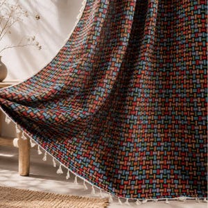 May include: A fabric curtain featuring a repeating woven pattern of small rectangular blocks in shades of red, blue, green, orange, and yellow on a black background, finished with a white fringe trim along the bottom edge of the textile.