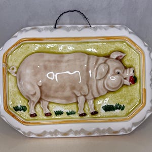 May include: A rectangular ceramic wall plaque featuring a raised, light brown pig standing on a textured light green background. The pig is eating a small red item. The plaque has a white scalloped border with orange trim and a black wire hanger.