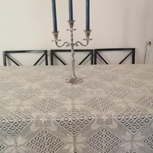 Può includere: Un candelabro in metallo color argento a tre bracci con candele affusolate blu scuro, posizionato su una tovaglia rettangolare bianca caratterizzata da un intricato motivo a uncinetto ripetuto, bordo smerlato e motivi geometrici a stella.
