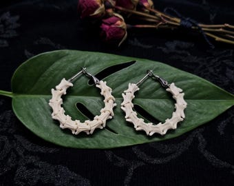 May include: A pair of hoop earrings constructed from small, off-white, bone-like vertebrae segments arranged in a circular shape. Each earring features a silver-toned metal clasp mechanism at the top for securing the hoop in place.