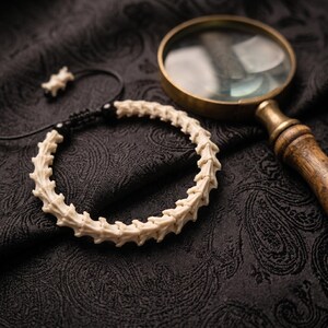 May include: A bracelet constructed from a series of off-white, bone-like vertebrae segments strung together on a black cord with a sliding knot closure, accompanied by a magnifying glass with a wooden handle and a brass-toned metal frame.