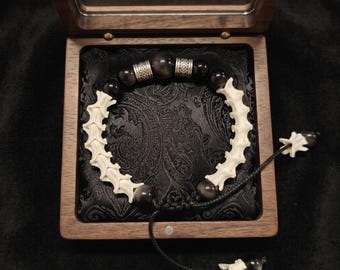 May include: A bracelet featuring alternating segments of off-white bone-like vertebrae and dark, polished obsidian-style beads. The design includes two silver-toned metal spacers with intricate patterns and a black cord with an adjustable closure.