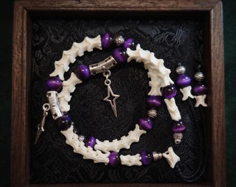 May include: A necklace featuring alternating white bone-like segments and round purple cat's eye beads, accented with silver-toned metal spacers and two star-shaped charms, presented inside a dark wooden box with a black fabric lining.