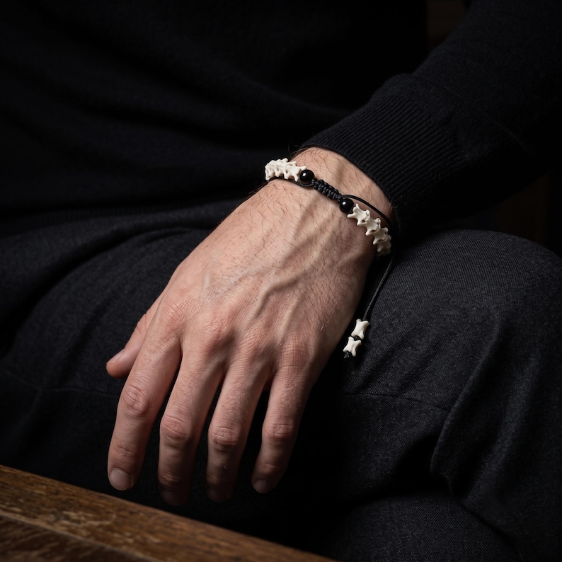 May include: A bracelet featuring a series of off-white, bone-like vertebrae segments alternating with smooth, round black beads. The components are strung on a black cord with an adjustable sliding knot closure and two small bone-like beads at the ends.