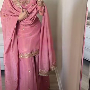 May include: A dusty pink salwar kameez set featuring a long-sleeved tunic and wide-leg trousers. The garment is adorned with intricate gold embroidery, metallic thread work, and small reflective embellishments along the neckline, cuffs, and hem.