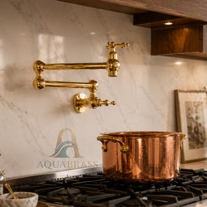 May include: A polished gold wall-mounted pot filler faucet with a double-jointed swing arm and cross-handle controls, positioned above a black gas stovetop featuring a large copper pot. The logo AQUABRASS is visible on the marble backsplash.