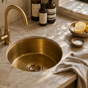 May include: A round, brushed brass kitchen sink set into a light beige stone countertop. A matching brass gooseneck faucet is positioned to the left. Nearby are two dark glass bottles, a wooden bowl with sliced lemons, and a small bowl of salt.