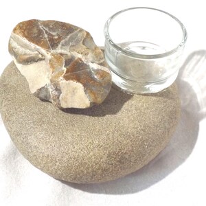 May include: A natural stone candle holder with a clear glass votive cup. The holder is composed of two stones: a large, rounded, light brown stone and a smaller, multi-colored stone. The votive cup is empty and sits on top of the larger stone.