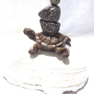May include: A bronze-colored turtle figurine with a stack of gray stones balanced on its back. The turtle is positioned on a white, textured seashell, creating a naturalistic decorative piece.