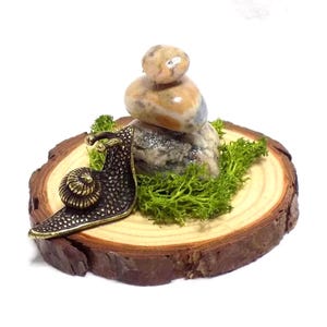 May include: A bronze-colored snail figurine on a wooden slice, with a small stack of stones and green moss. The snail has a detailed shell and textured body. The wooden slice has a natural, rustic appearance.