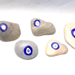 May include: Five painted stones with a blue and white eye design. The stones vary in shape and color, including gray, beige, and off-white. The eye design features a blue iris and a white center.