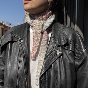 May include: A hand-crocheted neck scarf featuring a variegated color palette of soft pink, cream, and light blue tones. The scarf has a textured, ribbed stitch pattern and is tied in a simple knot at the front, worn over a white shirt and a black leather jacket.