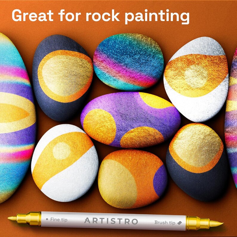 May include: A white dual-tip marker with gold caps labeled ARTISTRO, featuring a fine tip and a brush tip. The marker is positioned below several smooth, rounded stones decorated with vibrant, multi-colored metallic paint designs.