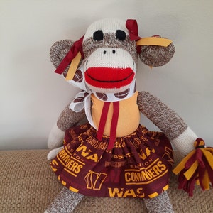 May include: A sock monkey wearing a burgundy and gold skirt with the words "Washington Commanders" printed on it. The monkey has a red mouth, white face, and brown and white striped socks.
