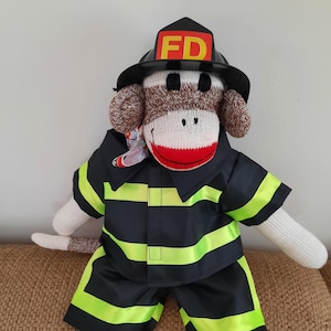 Puede incluir: Un muñeco de mono de calcetín hecho a mano vestido de bombero. El mono lleva un casco negro con "FD" amarillo, una chaqueta y pantalones cortos negros con rayas amarillo neón. La cara del mono es blanca con una boca roja.
