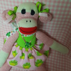 May include: A handmade sock monkey doll with a pink and white color scheme. The doll has a green dress with a frog print, a red mouth, and green bows. The doll is made of socks and fabric.