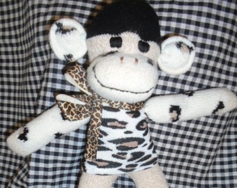 Puede incluir: Un juguete de peluche de mono con pelo negro, ojos marrones y una camisa a rayas blancas y marrones. El mono está sentado sobre una tela a cuadros blanco y negro.