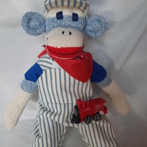 May include: A sock monkey doll wearing a blue and white striped engineer cap and matching overalls over a blue shirt. The doll has a red bandana around its neck and a small red plastic toy train attached to the front of the striped overalls.