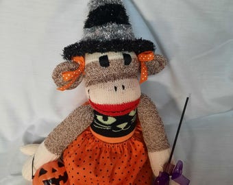 Puede incluir: Un muñeco de calcetín con forma de mono disfrazado de bruja, con un sombrero de espumillón negro y plateado con cintas naranjas de lunares, una falda naranja con puntos negros y un top negro con una cara de gato verde. Sostiene una calabaza.