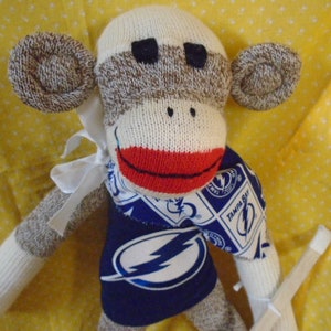 May include: A sock monkey wearing a blue and white jersey with a lightning bolt logo and the text "Tampa Bay". The monkey has a red mouth and brown eyes.