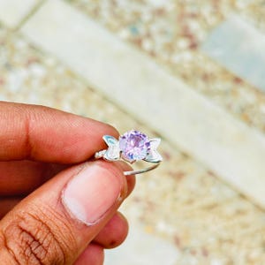 May include: A silver ring featuring a round, faceted light purple gemstone at the center. The setting includes two small, wing-shaped silver accents on either side of the stone, creating a symmetrical design on a thin, polished silver band.