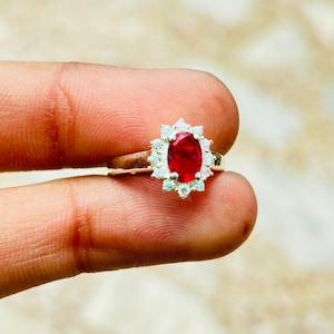 Può includere: Un anello in argento con una gemma centrale rosso intenso dal taglio ovale, circondata da un alone di dodici piccole pietre chiare dal taglio rotondo. La fascia in metallo è lucida e liscia, con un classico design a grappolo centrale.