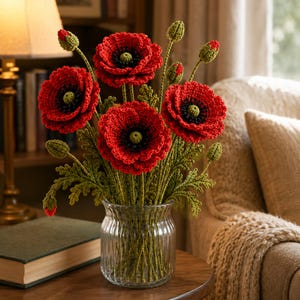 May include: A bouquet of crocheted red poppy flowers with dark centers and green stems, leaves, and buds arranged in a clear, vertically ribbed glass vase. The flowers feature textured petals and detailed centers, standing upright in the transparent container.