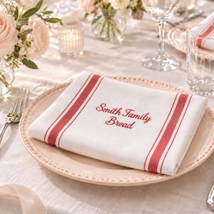May include: A white rectangular cloth napkin featuring two parallel red stripes along each side and the embroidered red text Smith Family Bread centered on the fabric, resting on a light pink plate with a beaded edge.