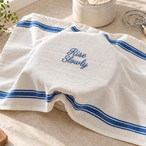 May include: A white kitchen towel featuring two horizontal blue stripes near the top and bottom edges. The center of the towel is embroidered with the blue text Rise Slowly in a cursive script font, draped over a round object.