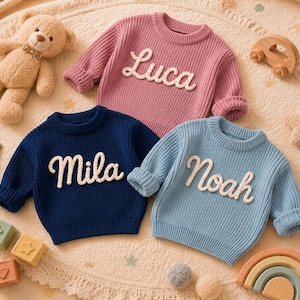 May include: Three long-sleeve knit sweaters in pink, navy blue, and light blue. Each sweater features a white embroidered name on the front: Luca, Mila, and Noah. The sweaters are displayed alongside a teddy bear and various wooden toys.