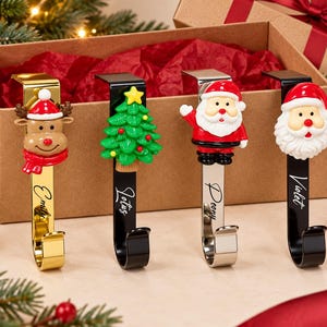 May include: Four metal stocking holders with holiday-themed figures: a reindeer, a Christmas tree, and two Santa Clauses. Each hook features a personalized name engraved in script: Emily, Lotus, Peony, and Violet. The hooks have gold, black, and silver finishes.