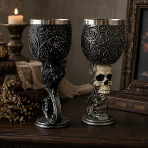 May include: Two decorative goblets with stainless steel inserts. The left goblet features a black cat base and a pentagram emblem. The right goblet features a human skull base. Both have ornate, dark, swirling relief patterns on the bowl.
