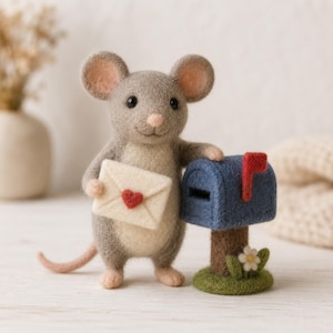 Puede incluir: Un ratón de lana fieltrada gris con vientre color crema, sosteniendo un sobre crema con un pequeño corazón rojo. El ratón está junto a un buzón azul con una bandera roja, sobre un poste marrón con una base verde y una pequeña flor blanca.
