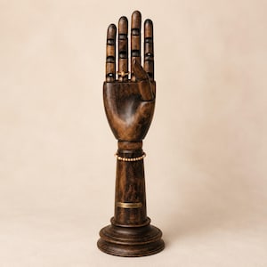 Op de afbeelding: Een donkerbruine houten gearticuleerde handdisplaystandaard met een ronde voet. De hand heeft beweegbare vingers, een goudkleurige ring aan de middelvinger, een goudkleurige kralenarmband om de pols en een klein rechthoekig messing naamplaatje.