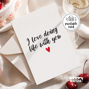 Może przedstawiać: Biała, fakturowana kartka okolicznościowa z czarnym napisem I love doing life with you oraz małym czerwonym sercem. Kartka leży na dopasowanej kopercie. Widoczna jest okrągła naklejka z ikoną drukarki i napisem printable card.