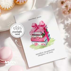 May include: A white greeting card featuring a watercolour illustration of a stack of books tied with a pink ribbon and decorated with small pink flowers. The text reads another year. and another wonderful chapter of you. on the card surface.